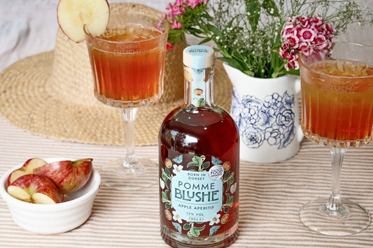 A bottle of Pomme Blushe apple aperitif centered on a table, surrounded by two filled glasses, apple slices, a small bowl of apples, and flowers in a vase. A straw hat is visible in the background.